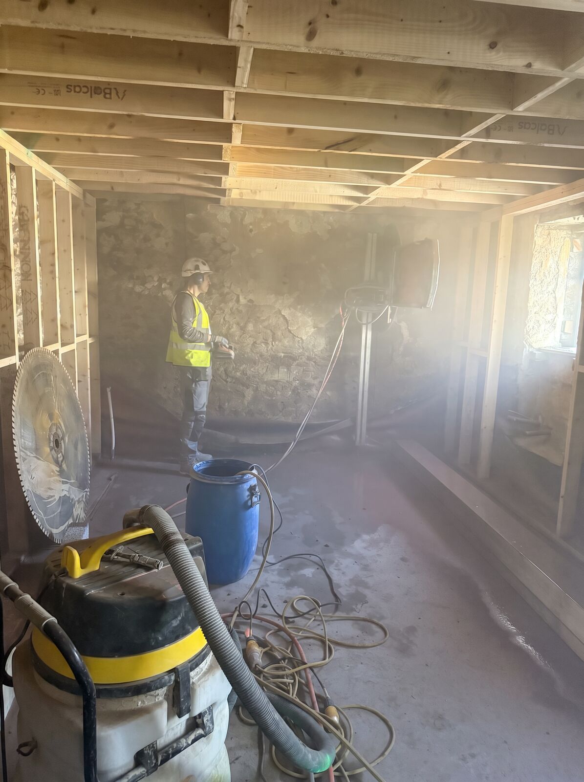 An operative running a track wall saw in a dust-filled room, with the large saw blade and site vacuum in shot