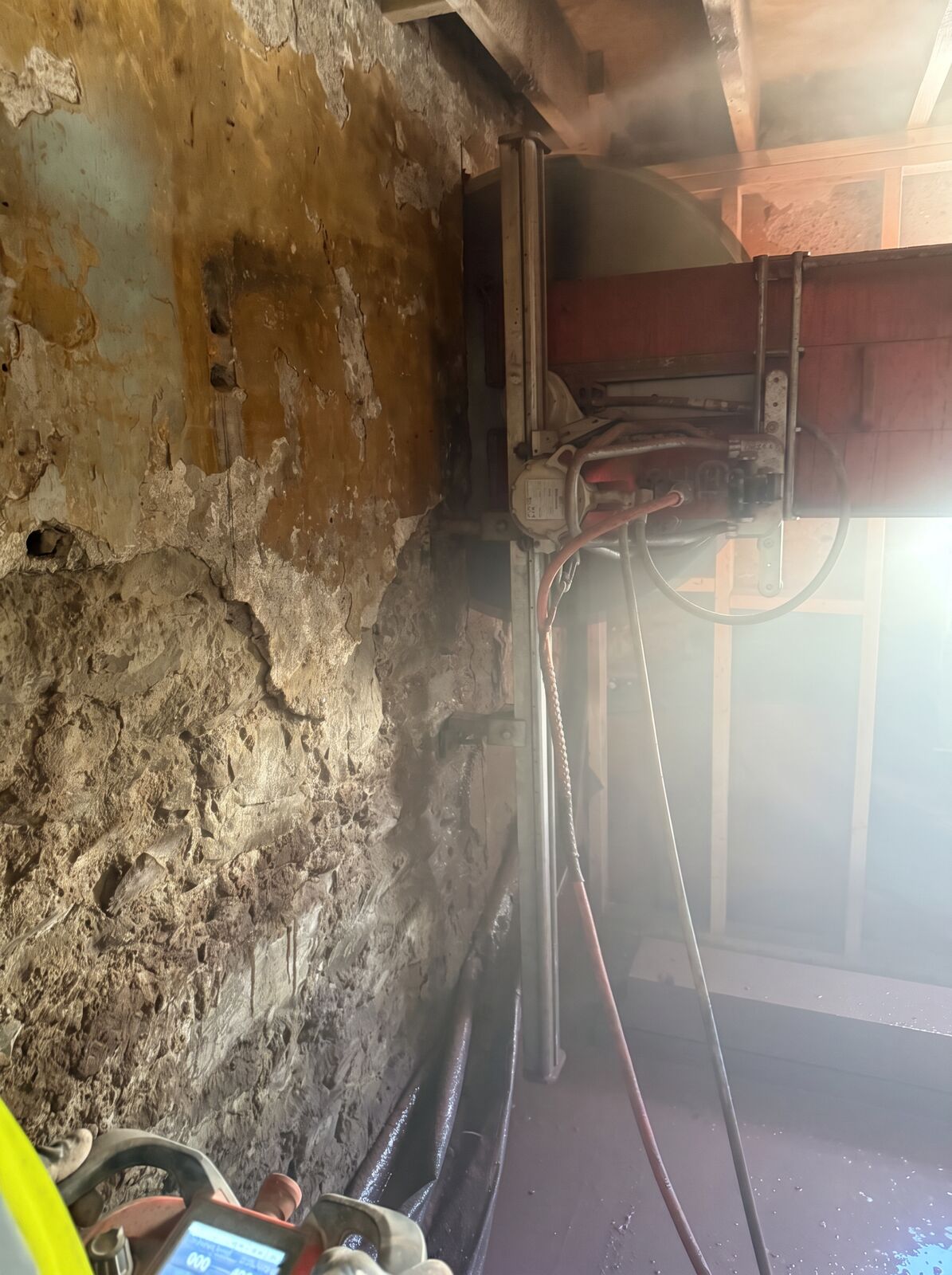 A vertical track wall saw cutting a reinforced concrete beam against an old wall on an interior job, water and dust in the air around the blade