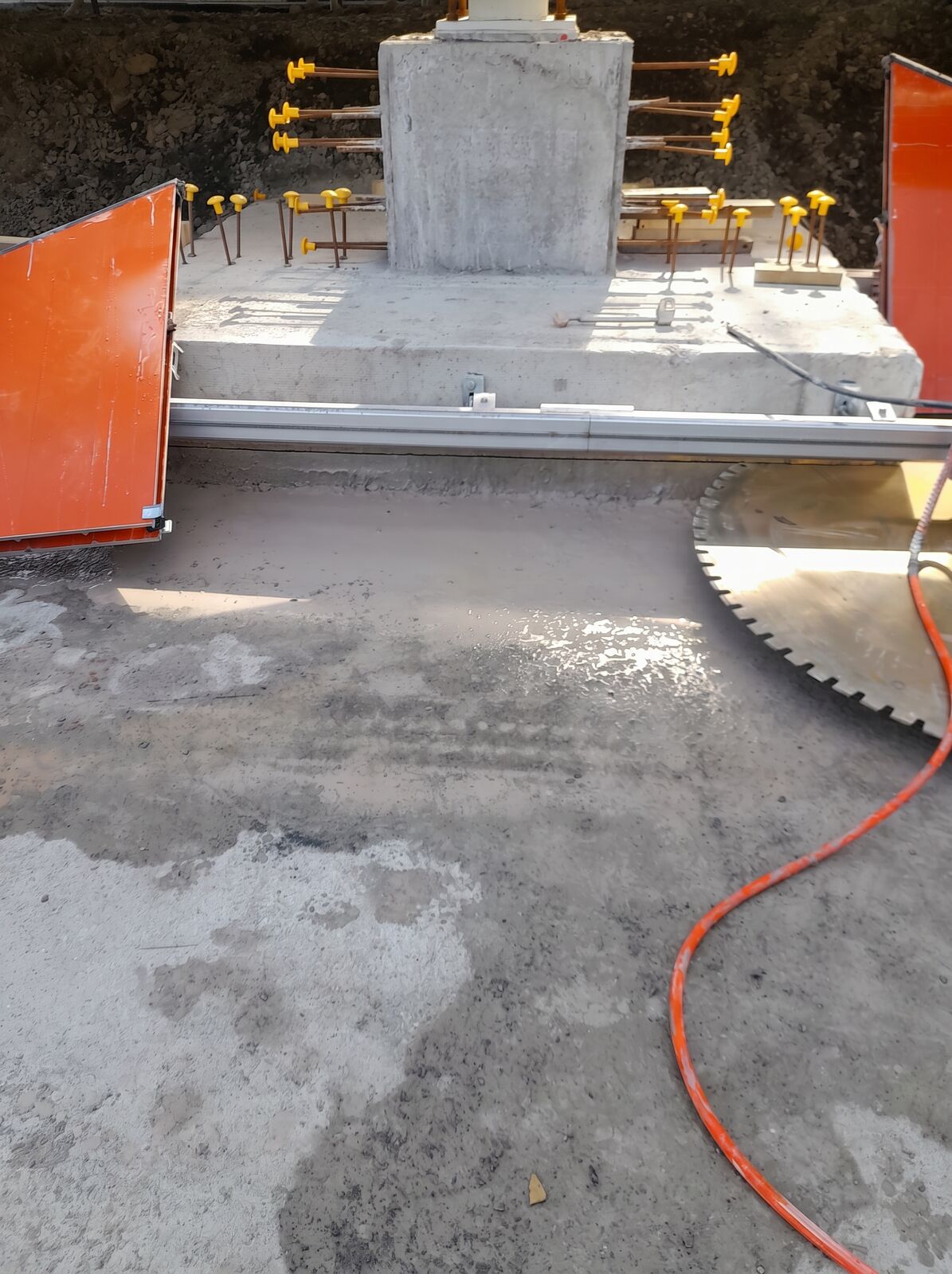 A track mounted wall saw set up around a concrete footing with the diamond blade lowered to the slab and yellow rebar caps marking the starter bars