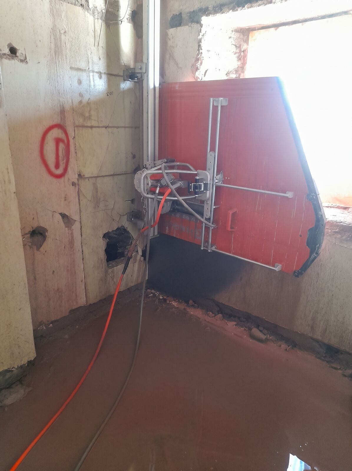 A red diamond rig anchored to a vertical track rail against a concrete wall, with water feed and power cabling running to it