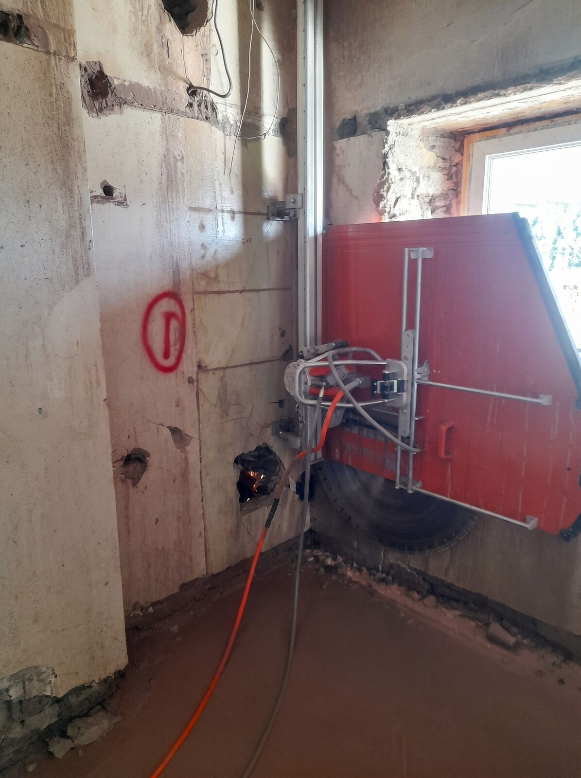 A red track-mounted wall saw cutting into a concrete wall beside a freshly opened window
