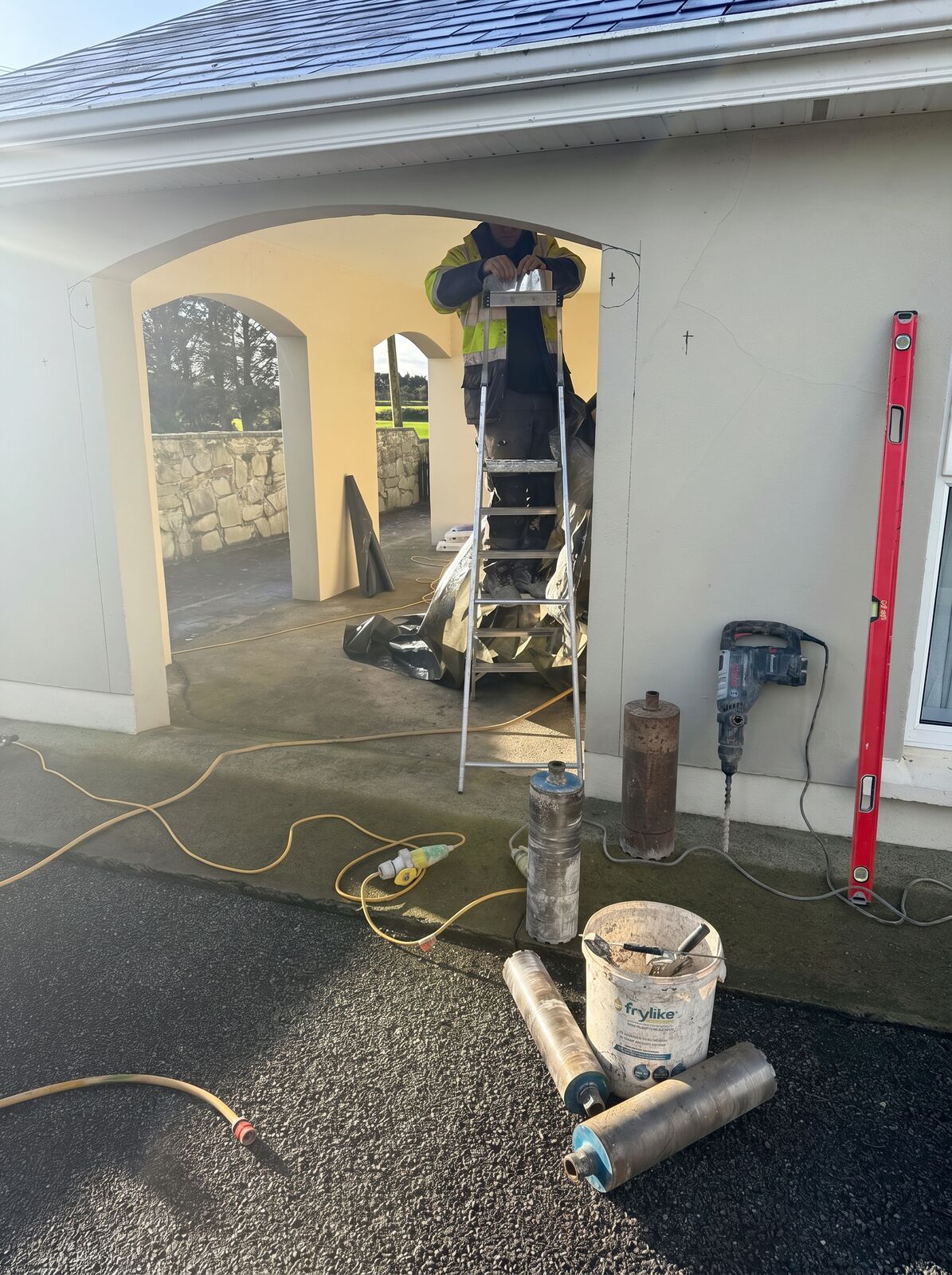 An operator up a stepladder core drilling through a house archway, with cut core barrels and a hammer drill on the ground