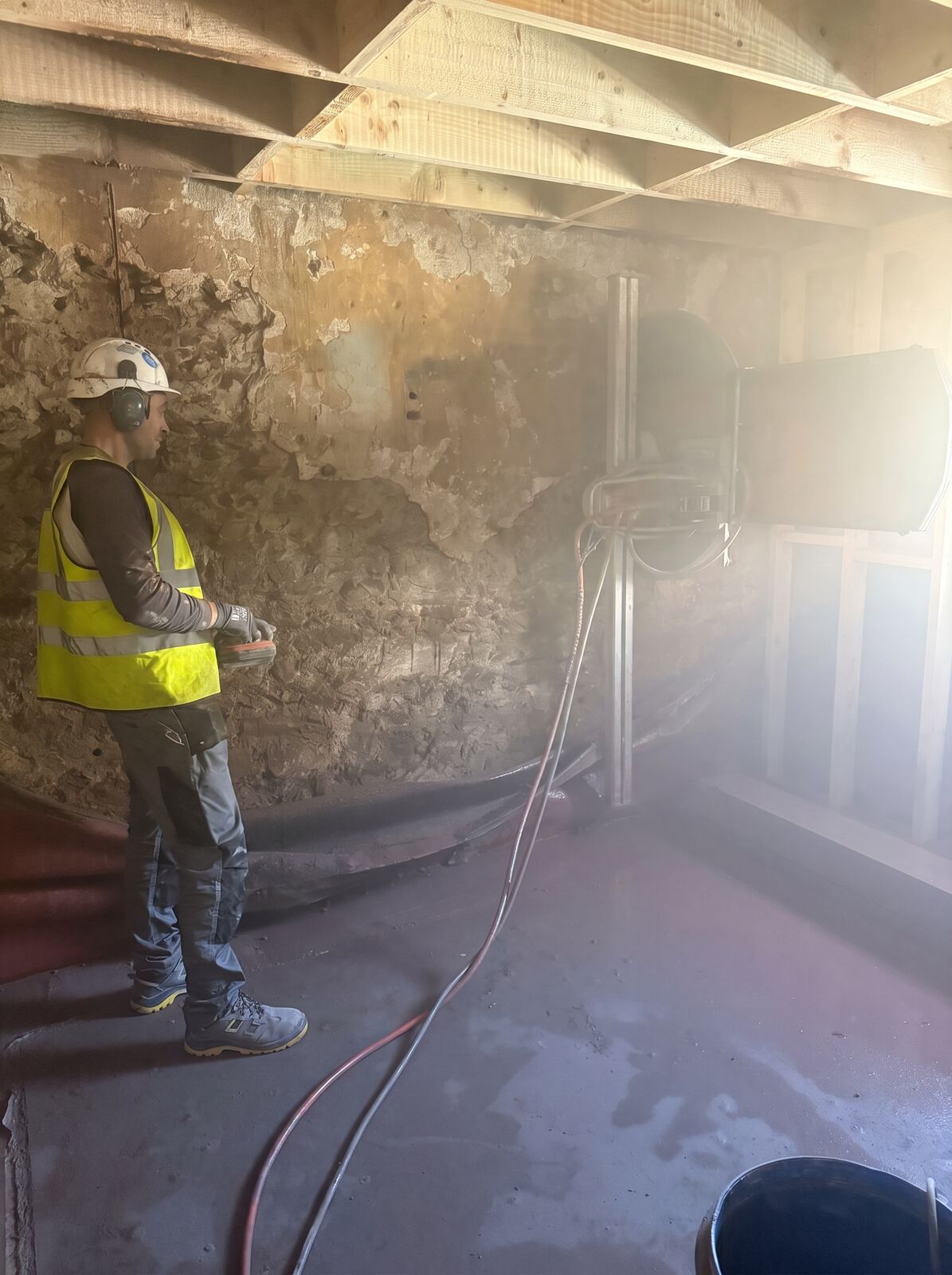 An operative running a track wall saw across an old stone wall in a dust-filled room