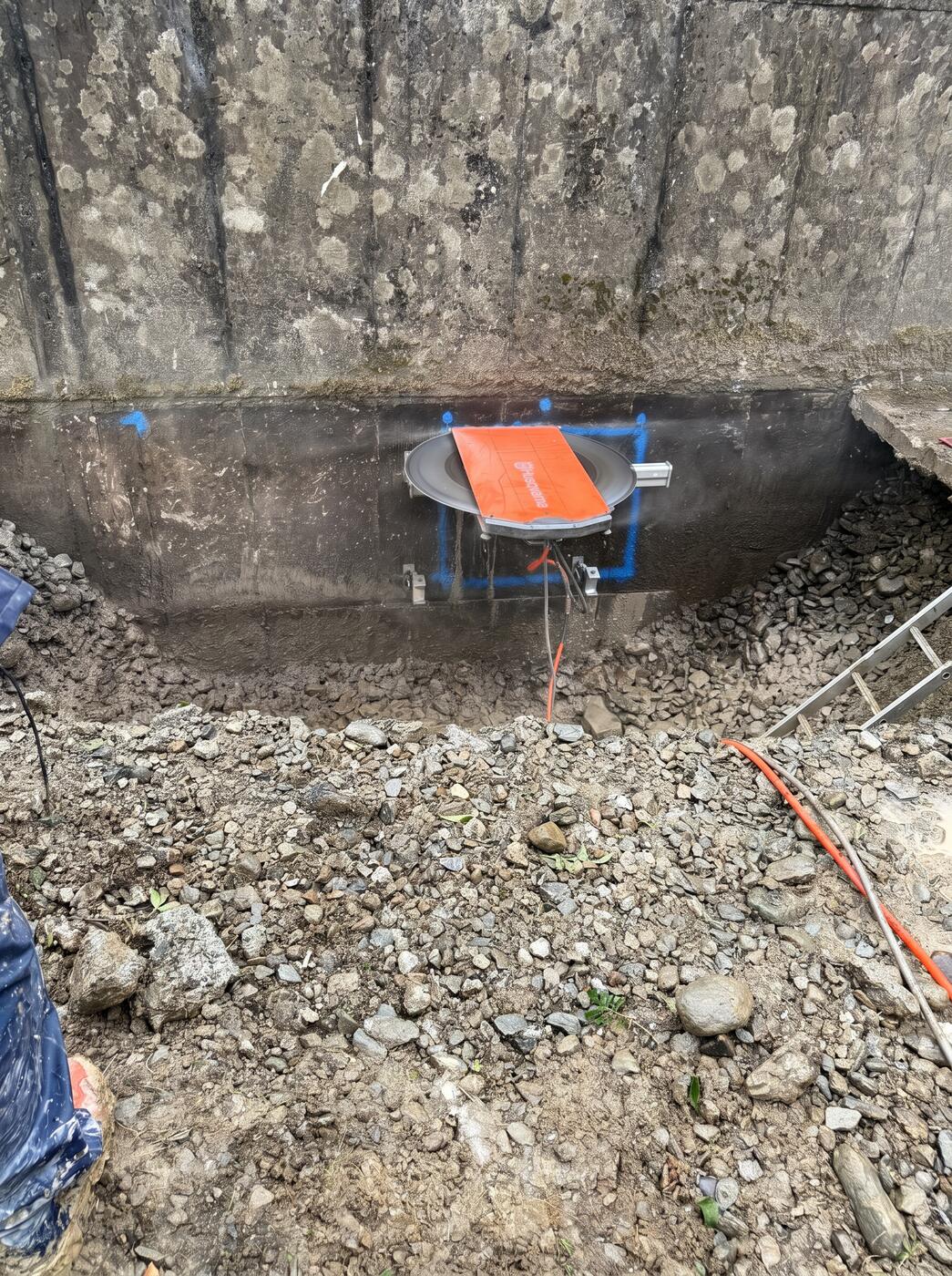 A Husqvarna saw cutting a horizontal line along the base of a concrete wall above an excavated rubble trench