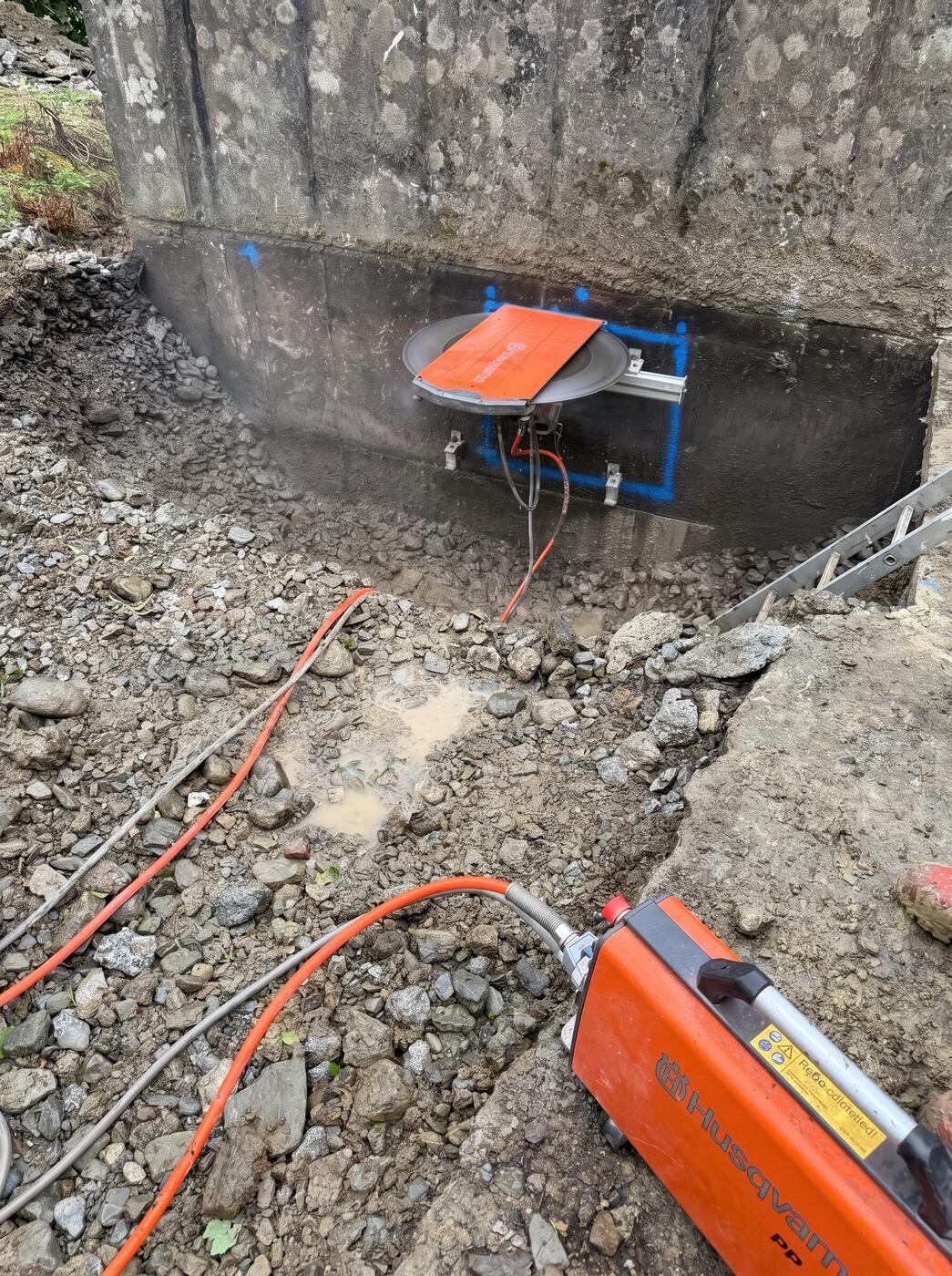 An orange Husqvarna track saw cutting into a concrete base, blue setting out marks around the blade, with the power pack and water feed running out across a rubble trench